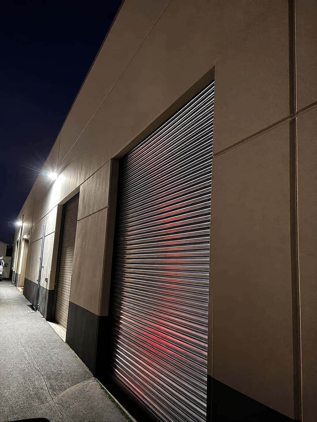 Two commercial roll up doors on an industrial building under a wall pack light at night