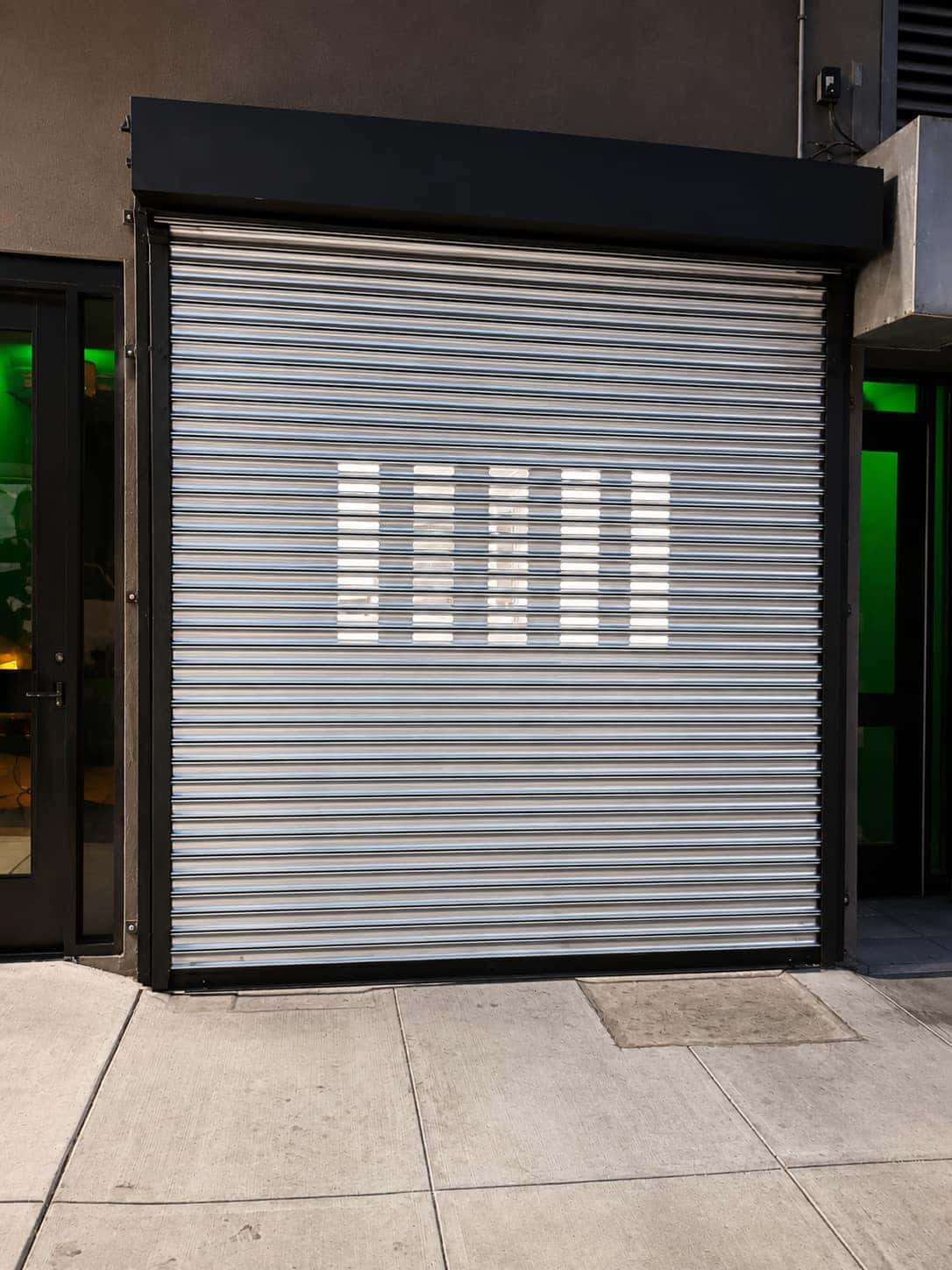 Perforated roll up security gate with lighted vision slots closed over a retail storefront
