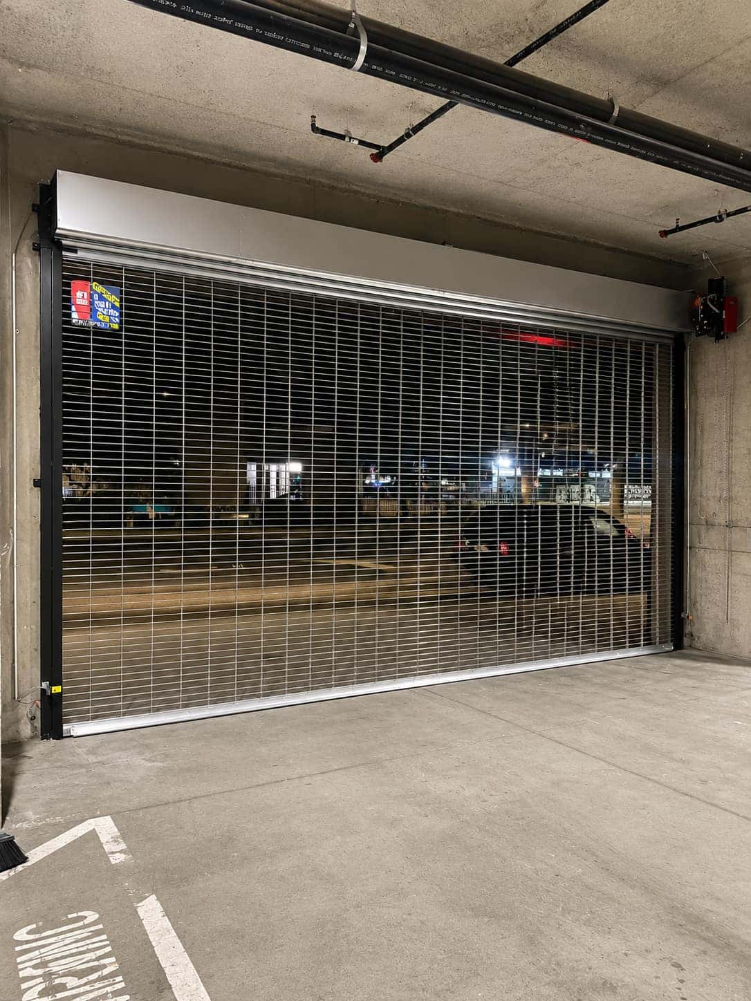 Open-air rolling security grille closed across a parking garage bay with a side-mounted operator